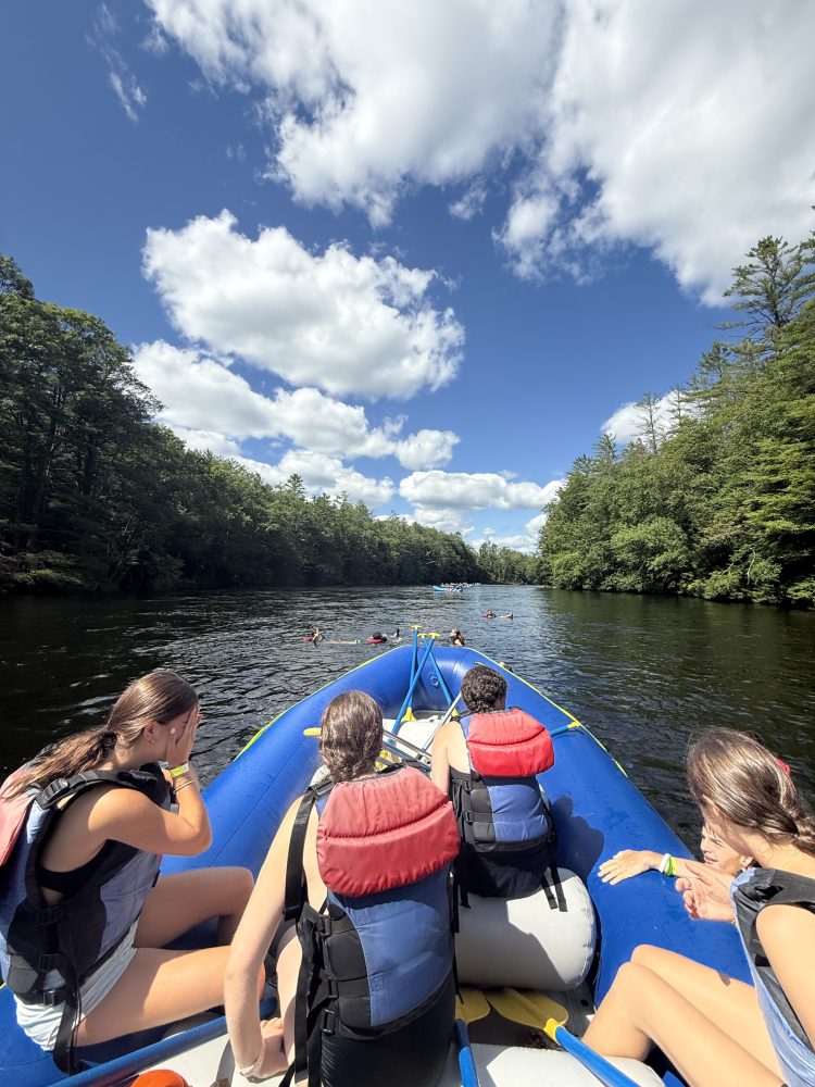 Four people in a raft on a river surrounded by trees on a sunny day.