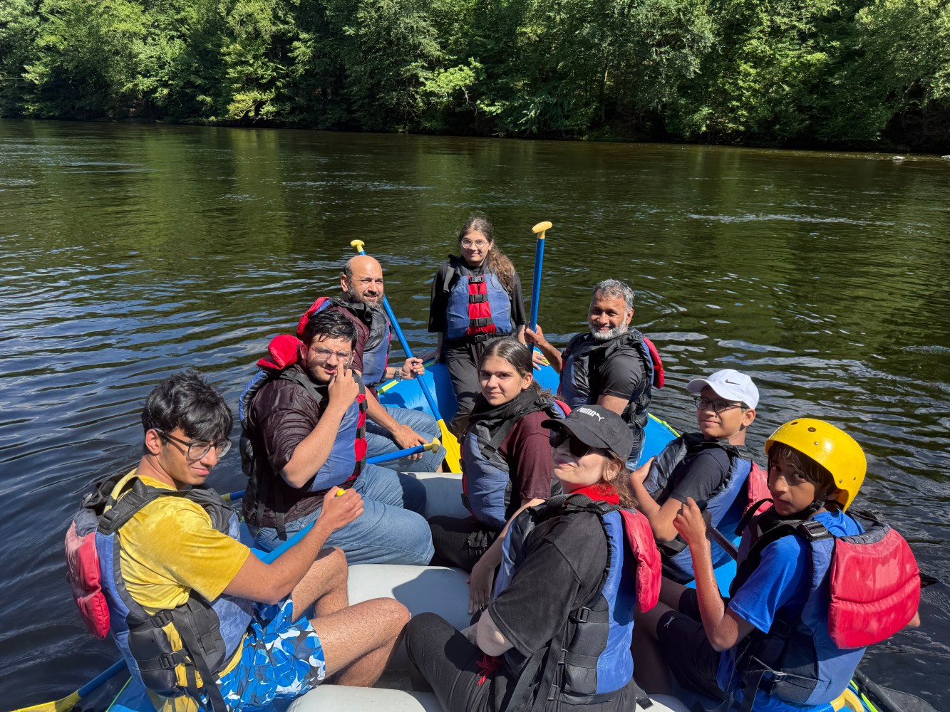 Group rafting on a river, wearing life jackets and helmets, surrounded by trees.
