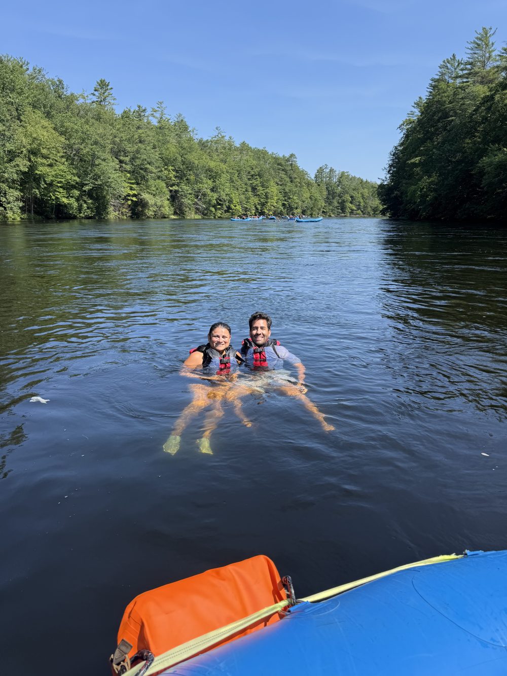 Two people in life vests floating on a lake beside a kayak.